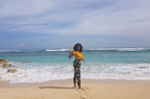 A person in a yellow top and colorful pants waves on the beach in Bali smiling